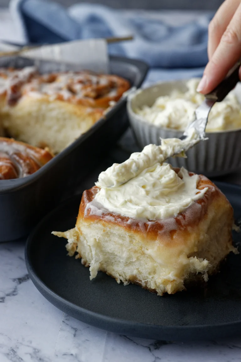 Hand spreading cream cheese frosting on cinnamon scroll