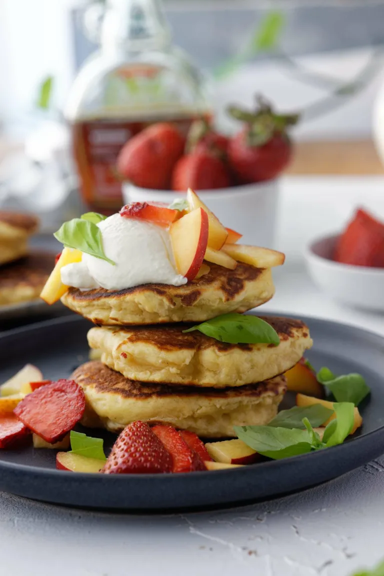 Stack of 3 pancakes on grey plate loaded with cream, strawberries, nectarine slices and basil leaves.
