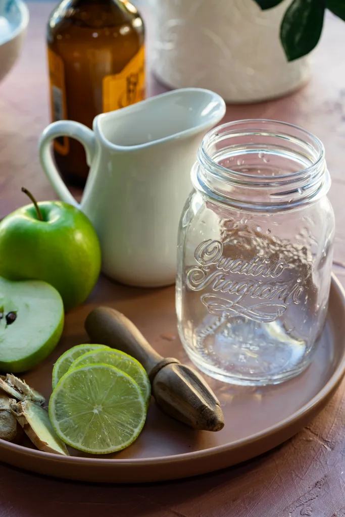 Glass mason jar on pink plate surrounded by lime slices, apples and ginger beer.