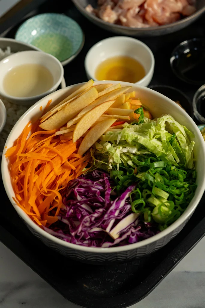 Grey bowl of ginger apple slaw ingredients separated to showing how to make the recipe. Small bowls of ingredients in background