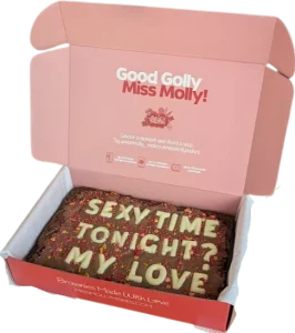 Slab of chocolate brownie in pink red box decorated with white chocolate letter and sprinkles with naughty phrasing.
