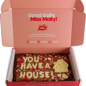 A large slab of chocolate brownie in a pink red box decorated with 'you have a house!' in white chocolate.